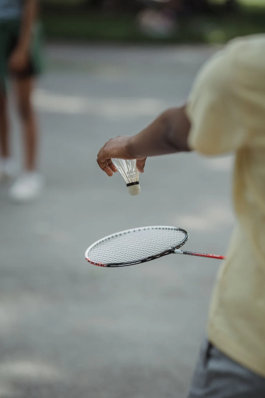 A hand holding a synthetic badminton shuttle beside a racket outdoors